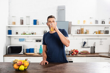  man drinking orange juice near bowl of ripe fruits on kitchen table