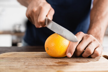 Cropped man holding orange and knife on wooden board on blurred background