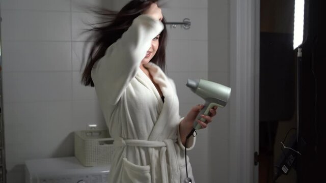 happy young woman drying hair using electric hair dryer and dancing in bathroom, having fun. Emotions and modern lifestyle concept.