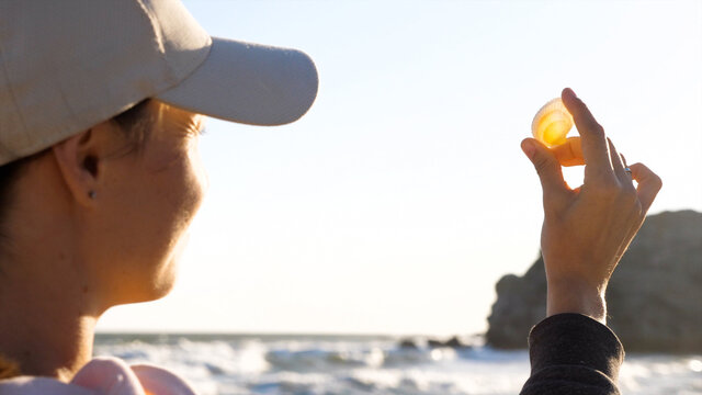 Young woman is holding seashell in hand and looking on sun through shell, close-up.