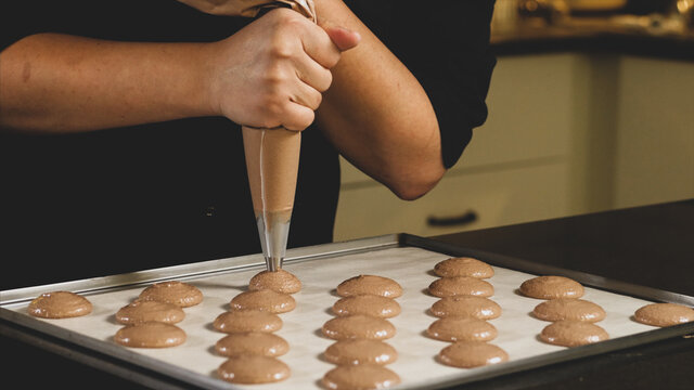 Chef making base for preparation macaron dessert. Close-up of extruding brawn creme from piping bag on metal tray for baking base of macaron dessert.