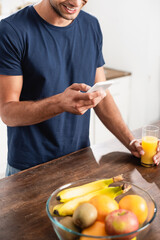 Cropped view of man using smartphone near glass of orange juice and fresh fruits on blurred foreground