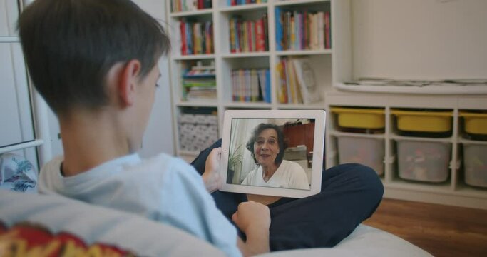 Grandparents Communicating Online with Their Grandchildren. Back View of Young Boy Sitting in his Room Having Online Conversation with his Grandmother via Digital Tablet.