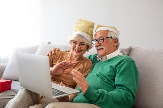 Happy senior couple in Santa hats sitting on couch at home and video calling family using laptop computer. Smiling grandparents keeping in touch with grandchildren on Christmas holidays in quarantine