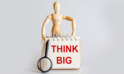 Wooden man shows with a hand to white notepad with text THINK BIG