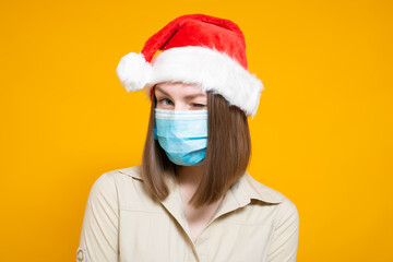Fototapeta na wymiar A beautiful young woman of Caucasian appearance with dark hair in a New Year's hat and a protective mask of the respiratory tract. On a yellow background.