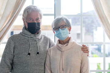 A smiling senior couple look at camera wearing surgical protective mask, in lockdown at home due to the coronavirus infection