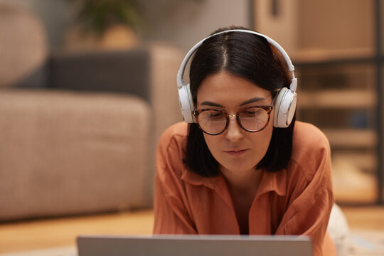 Portrait of contemporary woman working from home while lying on floor wearing headphones and using laptop, copy space