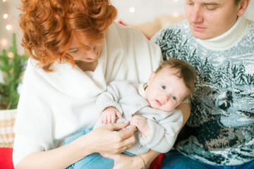 A young family in warm sweaters with a baby daughter in a Christmas bedroom interior decorated with red blankets, pillows, garlands and green pine needles. Family and Children. Christmas mood
