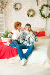 Obraz na płótnie Canvas A young family in warm sweaters with a baby daughter in a Christmas bedroom interior decorated with red blankets, pillows, garlands and green pine needles. Family and Children. Christmas mood 
