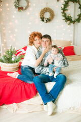 A young family in warm sweaters with a baby daughter in a Christmas bedroom interior decorated with red blankets, pillows, garlands and green pine needles. Family and Children. Christmas mood 