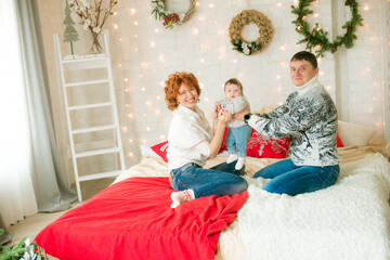 A young family in warm sweaters with a baby daughter in a Christmas bedroom interior decorated with red blankets, pillows, garlands and green pine needles. Family and Children. Christmas mood 