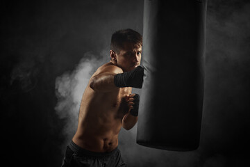 male boxer in black boxing wraps punching in boxing bag on dark background with smoke