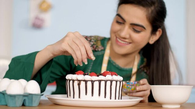 Young Indian chef decorating her freshly baked cake with confectionery sprinkles. Colorful sugar caramel sprinkled on a birthday cake for decoration - baking and decorating cakes concept