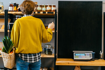 Minimalist vegan style girl with wicker bag and purchases on background of interior of zero waste shop. Woman in yellow shirt doing shopping without plastic packaging in plastic free grocery store.