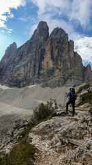 Fototapeta premium A man with big backpack hiking in high, Italian Dolomites. There are many sharp peaks behind. He is standing on a big boulder, enjoying the view. There are a few trees around. Sunny day. Outdoor