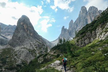 Fototapeta na wymiar A woman with big backpack and sticks, hiking in high Italian Dolomites. There are many sharp peaks in front of her. She is going up. There are a few trees around. Sunny day. Outdoor exercising