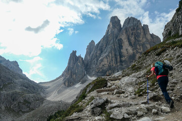 Fototapeta premium A woman with big backpack and sticks, hiking in high Italian Dolomites. There are many sharp peaks in front of her. She is going up. Lots of lose stones and landslides. Sunny day. Outdoor exercising