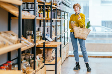 Minimalist vegan style girl with wicker bag and reusable glass coffee cup on background of interior of zero waste shop. Woman doing shopping without plastic packaging in plastic free grocery store. - obrazy, fototapety, plakaty
