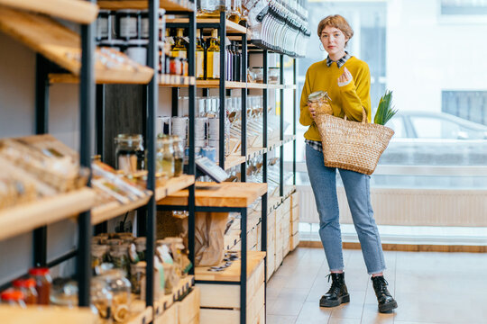 Minimalist vegan style girl with wicker bag and reusable glass coffee cup on background of interior of zero waste shop. Woman doing shopping without plastic packaging in plastic free grocery store.