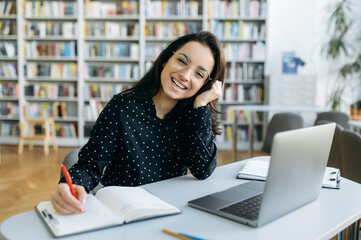 Portrait of an amazing caucasian young smiling woman, is sitting at the desk while remote work from home using laptop and looks into a camera