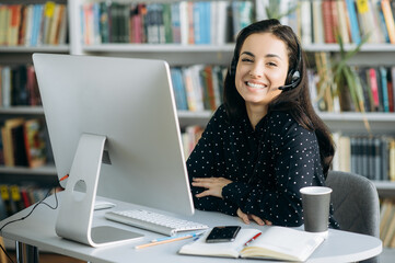 Remote work. Happy young pretty woman, call center worker, teacher or manager, wearing  a headset, looks at the camera with smile while sitting at her workplace - obrazy, fototapety, plakaty