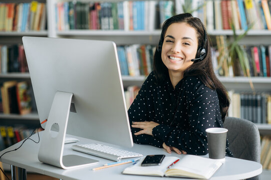 Remote work. Happy young pretty woman, call center worker, teacher or manager, wearing  a headset, looks at the camera with smile while sitting at her workplace