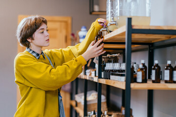 Young blond short haired woman filling natural biodegradable household chemical product from container dispenser in zero waste plastic free store. Dispensers for detergents, shampoo, soap, conditioner