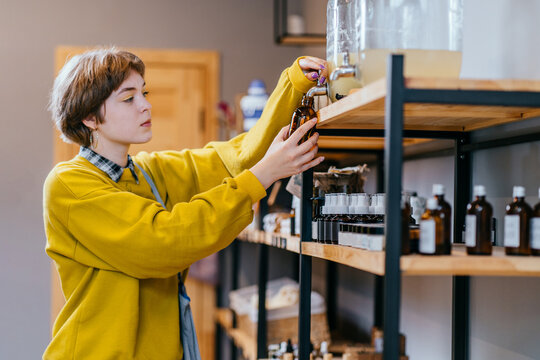 Young Blond Short Haired Woman Filling Natural Biodegradable Household Chemical Product From Container Dispenser In Zero Waste Plastic Free Store. Dispensers For Detergents, Shampoo, Soap, Conditioner