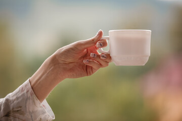 Morning coffee cup. Female hand holds white cup of morning hot drink - coffee or tea on the balcony on the background of mountain nature.