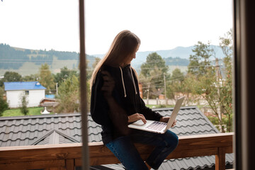 Closeup image of a woman working and typing on laptop while sitting on wooden balcony with green mountains on foggy day with blue sky background