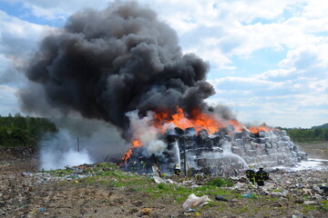 A group of firefighters extinguishes huge landfill fire with flames and black smoke in the background