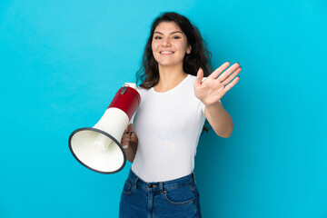 Teenager Russian girl isolated on blue background holding a megaphone and saluting with hand with happy expression