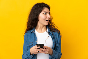 Teenager Russian girl isolated on yellow background using mobile phone and looking up