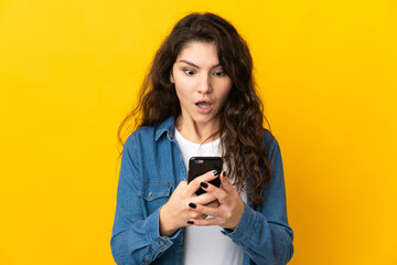 Teenager Russian girl isolated on yellow background looking at the camera while using the mobile with surprised expression