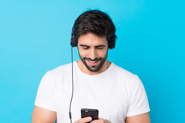 Young handsome man with beard over isolated blue background listening music and looking to mobile