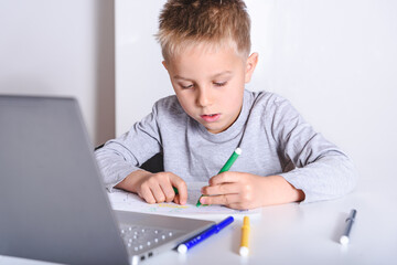 small kid drawing on a white table with a laptop near. Concept of home schooling during pandemic