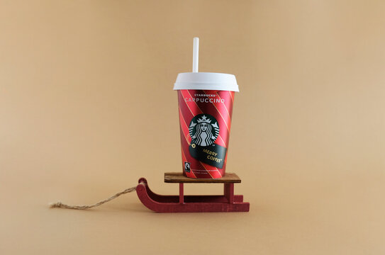 Disposable paper cup of coffee cappuccino with Starbucks logo on Decorative wooden red sleds. Colored pastel background. Seasonal winter Christmas coffee cup with a straw. MOSCOW - December, 2020.