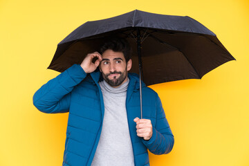 Man holding an umbrella over isolated yellow background having doubts and with confuse face...