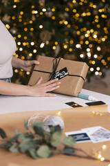 Woman Hands holding a Handmade Christmas Gift with a Hand Writing Christmas Card on Top, Tree in the Background and Wrapping Materials on the Table, Indoor, Evening, Close Up, Detail