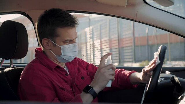 Man wearing mask in car desinfecting his hands to protect from coronavirus. Wearing protection masks man sanitizing stearing wheel during the second wawe coronavirus COVID19. Car Wash or carsharing