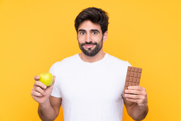 Young handsome man with beard over isolated yellow background taking a chocolate tablet in one hand...