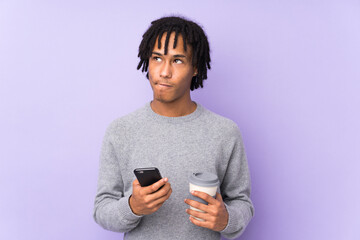 Young african american man isolated on purple background holding coffee to take away and a mobile...