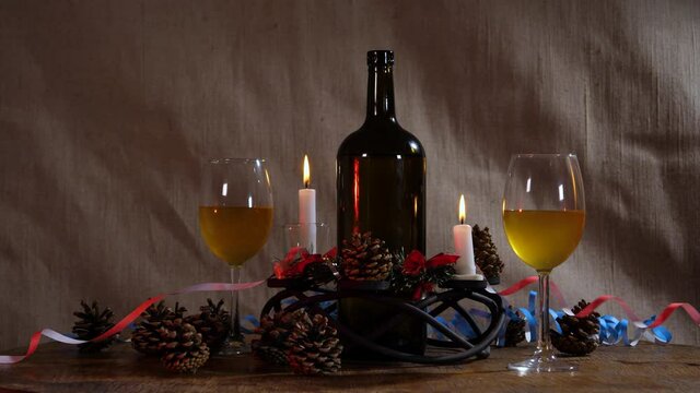 Against the background of decorative fabric there is a New Year's still life of lighted candles, glasses, a bottle of wine, Christmas cones, flying confetti and streamers.