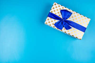 A gift wrapped in white paper with gold circles wrapped in a blue ribbon tied in a bow, isolated on a blue background, top view.