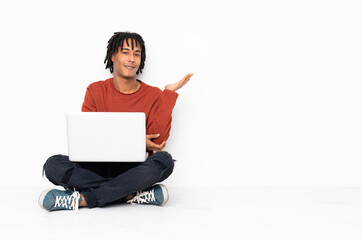 Young african american man sitting on the floor and working with his laptop extending hands to the side for inviting to come