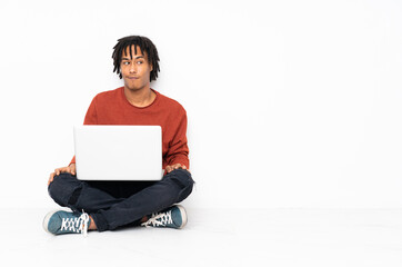 Young african american man sitting on the floor and working with his laptop making doubts gesture looking side