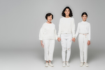 Smiling hisanic woman holding hands of daughter and mother on grey background, three generations of women