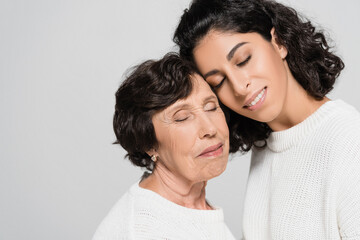 Smiling hispanic woman with closed eyes standing near senior mother isolated on grey, two generations of women
