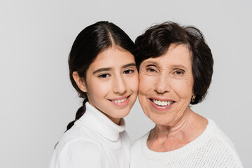 Senior woman smiling near hispanic granddaughter isolated on grey, two generations of women